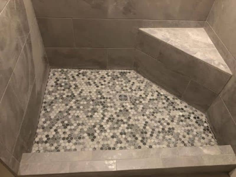 Custom tiled walk-in shower with gray wall tiles, mosaic hexagon floor design, and built-in corner bench - bathroom renovation