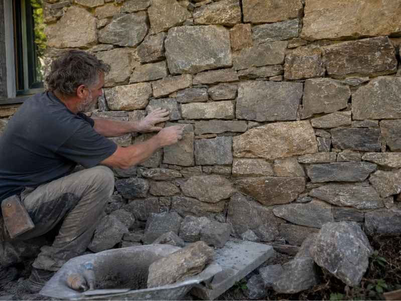 Stone mason blending natural stone Huntsville Alabama - Huntsville Brick Stone Man kneels beside a rough stone wall, placing rocks to build a dry-stone wall by a windowed building—construction scene.