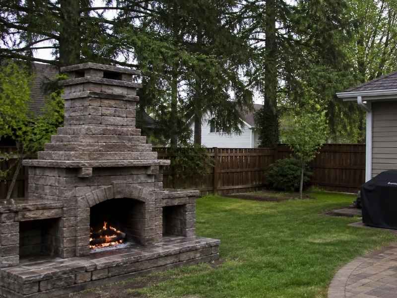 Outdoor Stone Fireplace Wind Exposed Yard Huntsville AL - Huntsville Brick Stone Outdoor stone fireplace in a residential backyard in Huntsville AL with open lawn, fencing, and surrounding trees.
